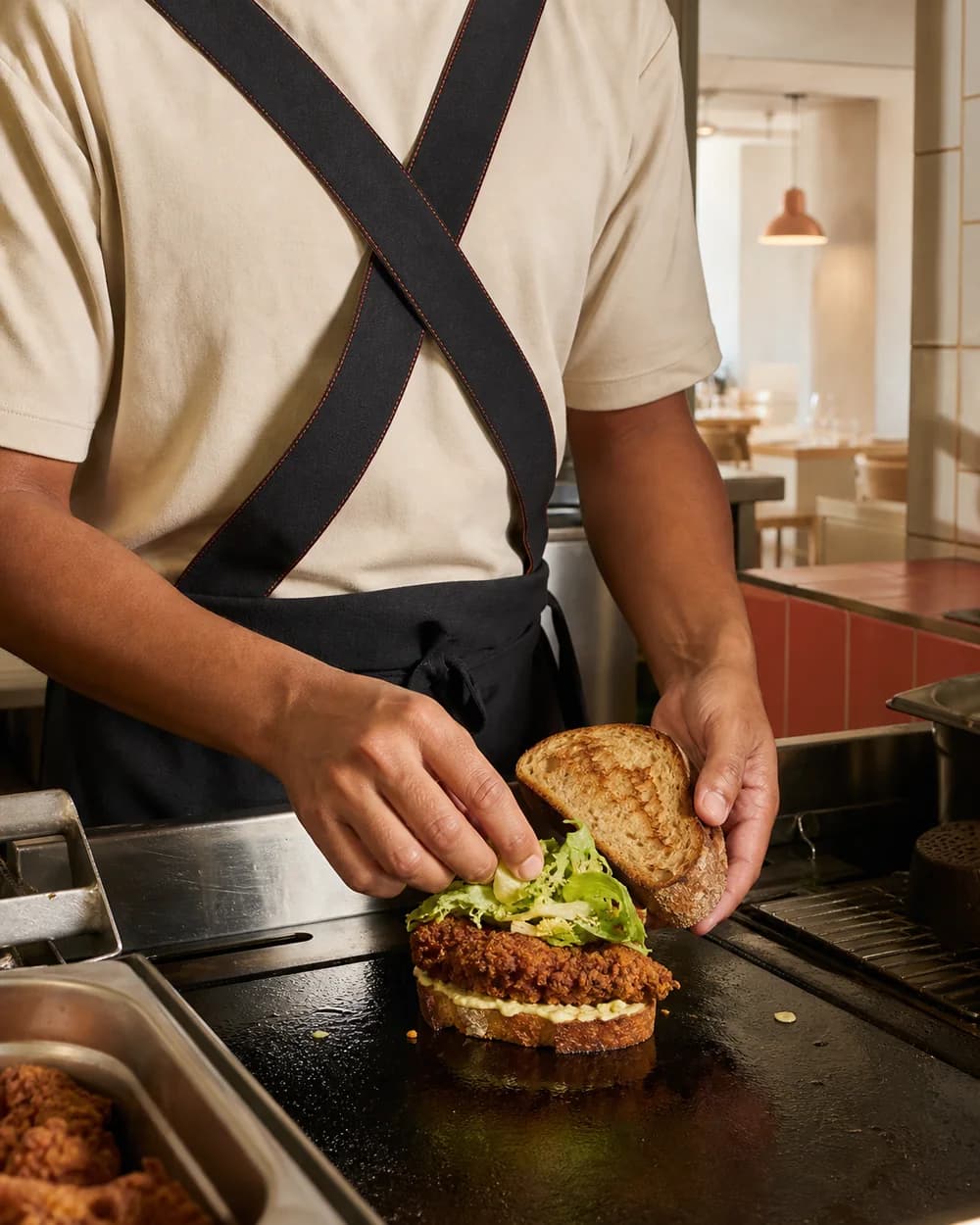 Cuisinier en tablier noir assemblant un sandwich au poulet croustillant dans la cuisine ouverte.