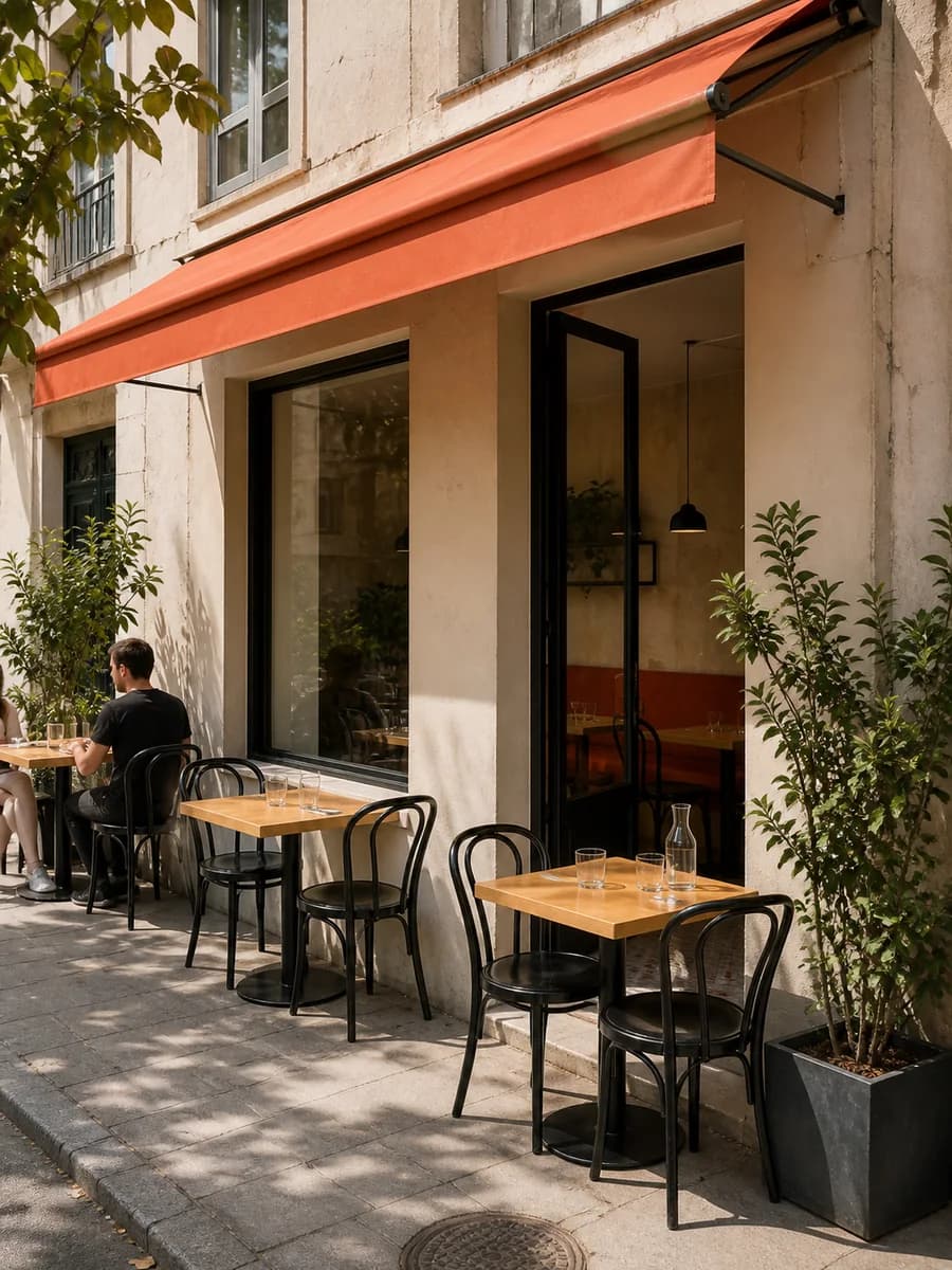 Terrasse de quartier sous un store corail, avec tables en bois et chaises en métal noir.