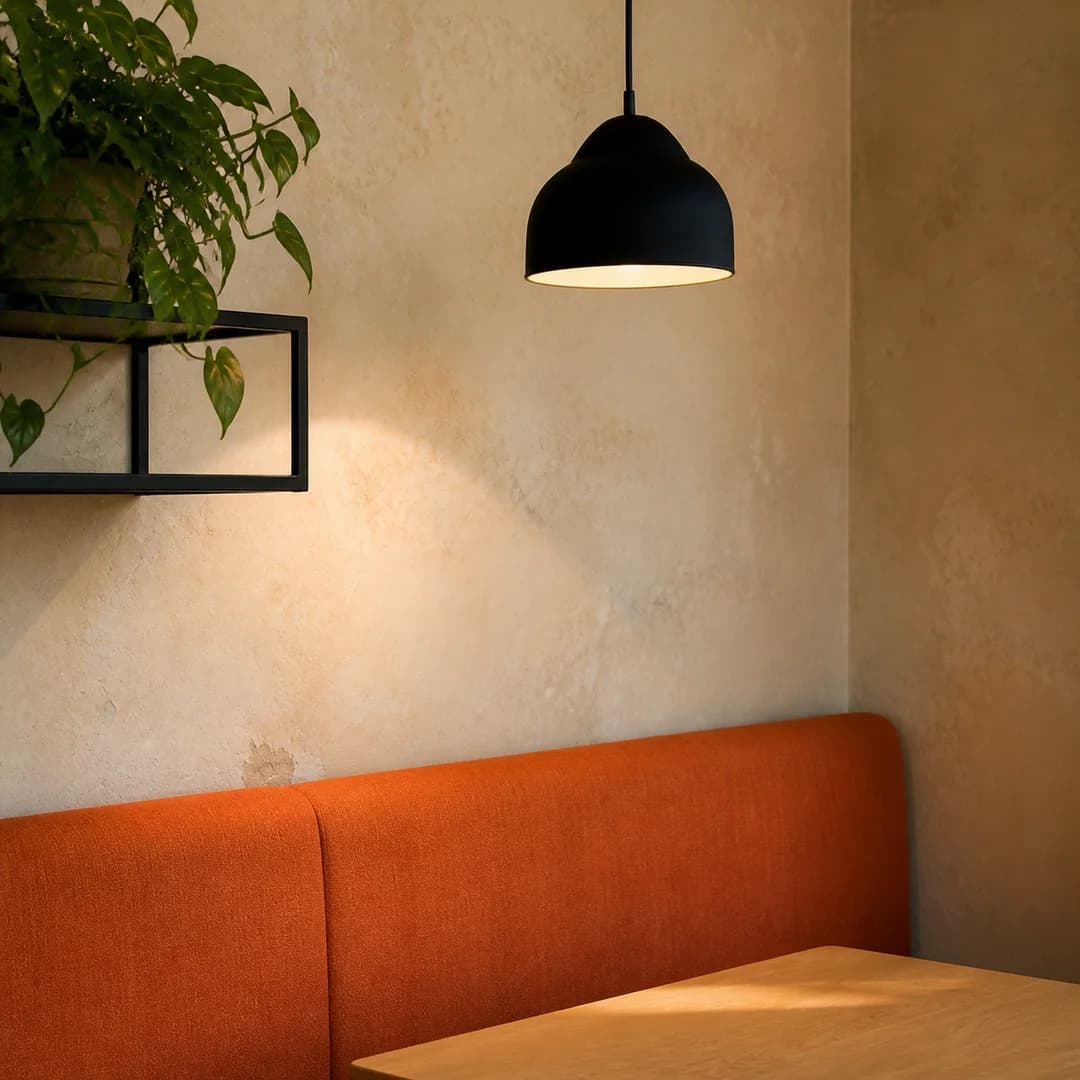 Coin de salle avec banquette corail, mur crème patiné, plante verte et suspension noire.